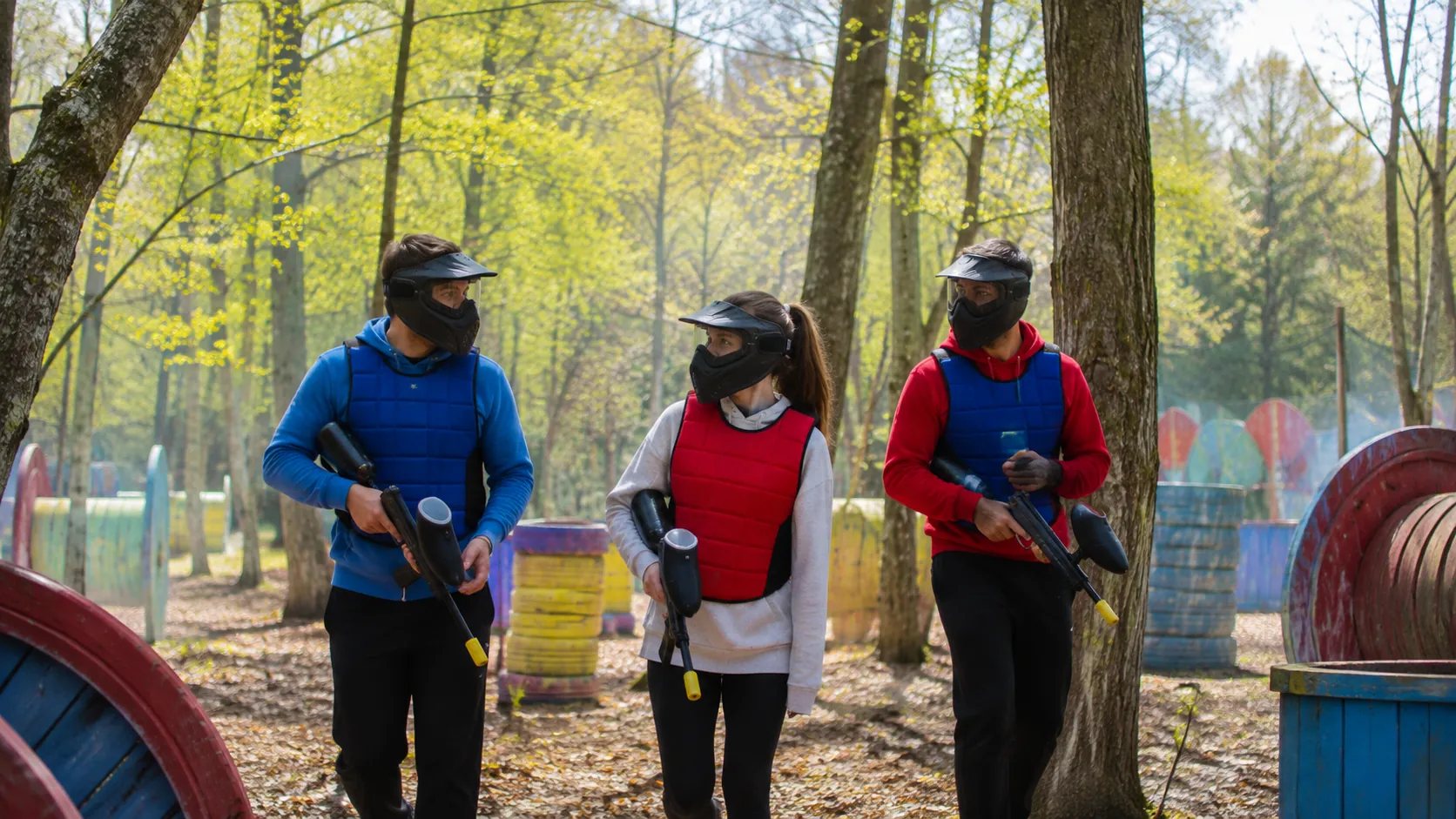 Trois amis équipés jouent au paintball au printemps sur un terrain boisé et lumineux