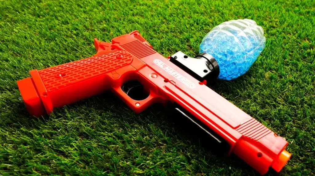 Lanceur Gel Blaster rouge posé dans l’herbe à côté d’un sachet de billes de gel bleues