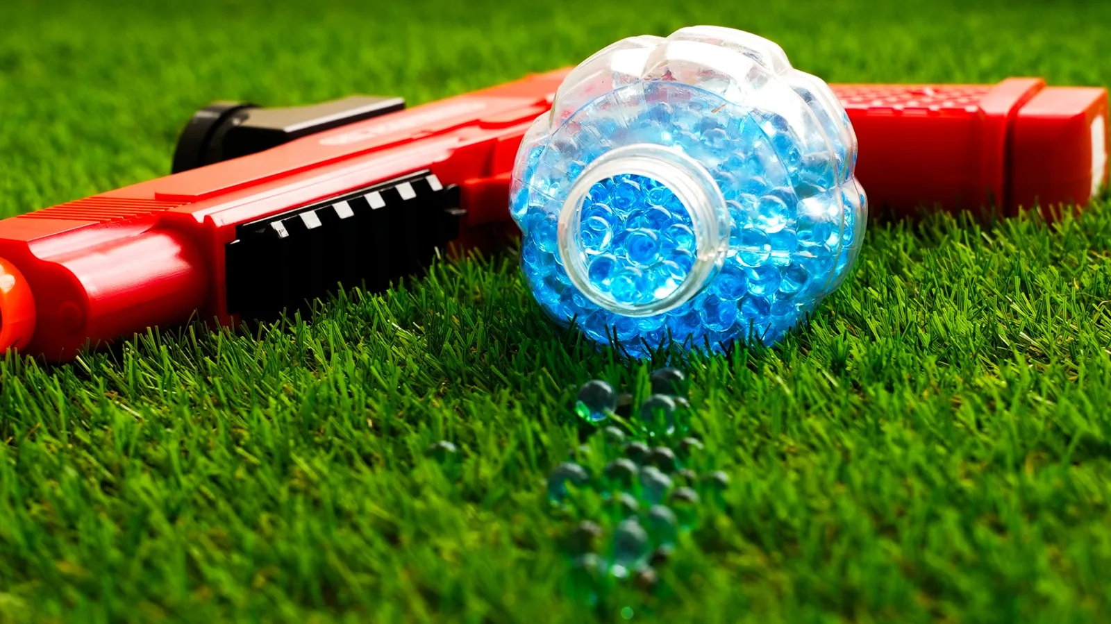 Billes de gel bleues versées à côté d'un lanceur Gel Blaster rouge sur gazon synthétique